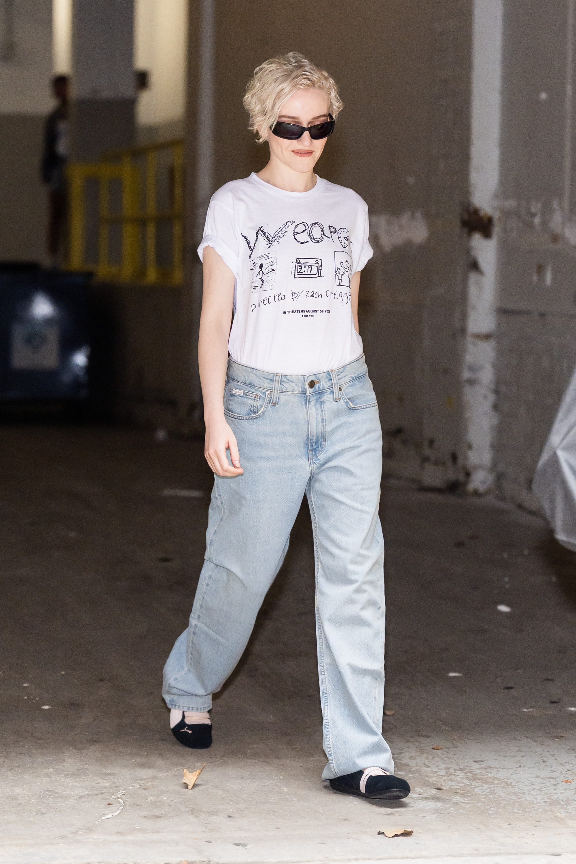 Julia Garner wearing a white graphic T-shirt with baggy jeans and black Puma Speedcat sneakers
