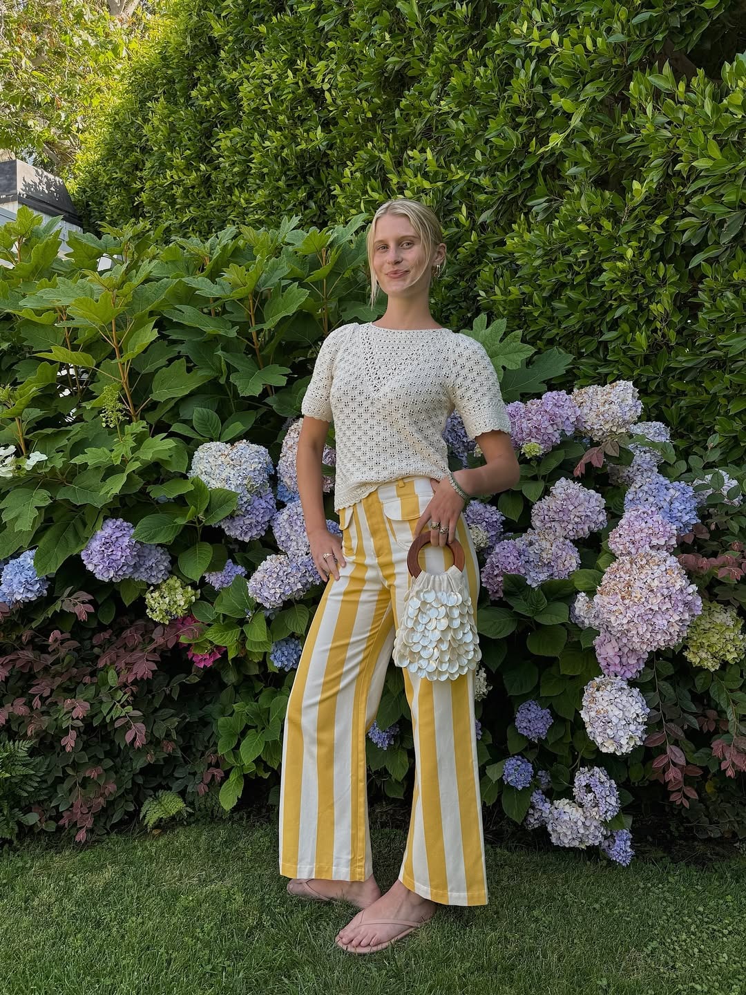 Influencer Lauren wears yellow and white striped cotton pants with a crochet white sweater and beige flip flops.