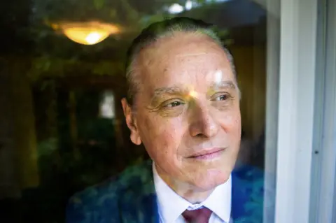 Dr Marrero, in a blue suit coat, white shirt and red tie, looks out from behind the window glass at his home in New Brunswick.
